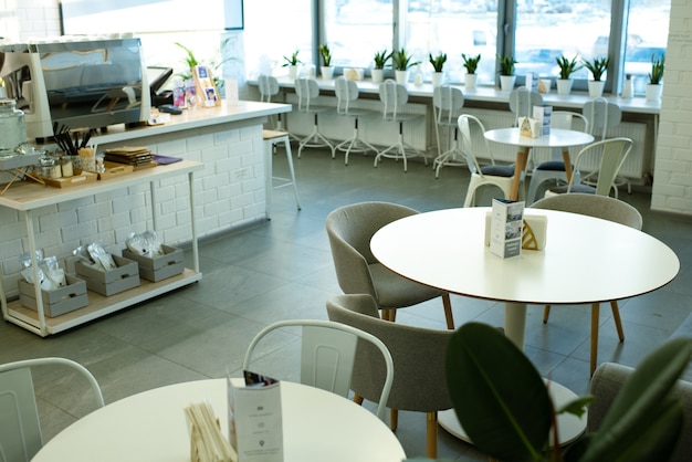 White round tables surrounded by comfortable armchairs and chairs along window inside cozy cafe