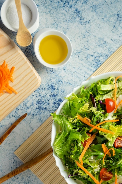 White salad bowl full of vegetables together with some ingredients. Oil, salt and tomatoes.