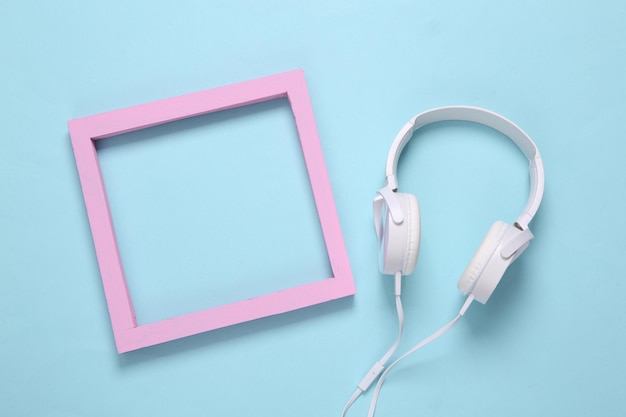 White stereo headphones and pink blank frame on a light blue background Copy space