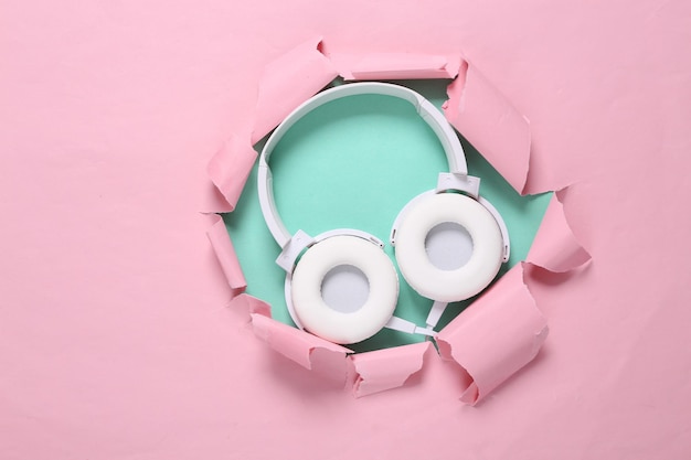 White stereo headphones through a torn hole on a bluepink background Minimalistic fashion still life