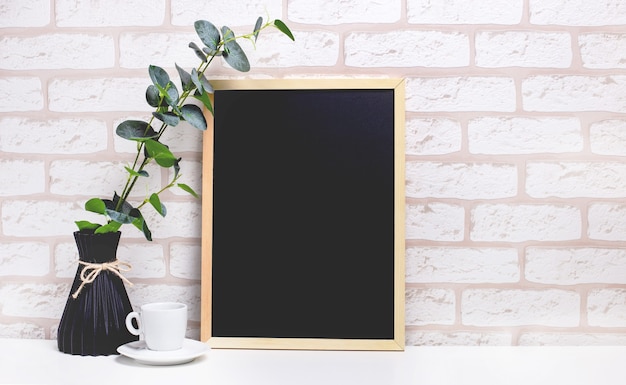 On a white table against a background of a light brick wall, a branch of eucalyptus in a dark vase, a white cup and a light wooden frame with a place to insert text. Home office. Copy space