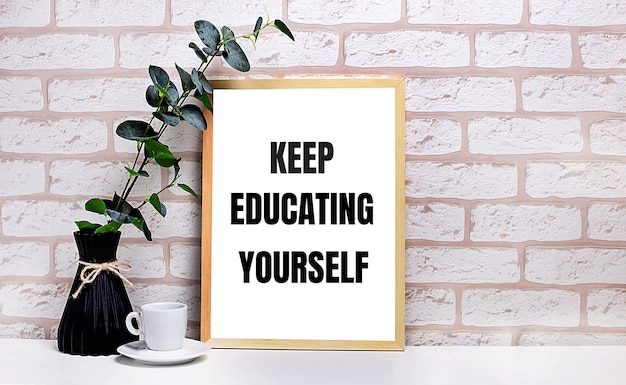 On a white table against a light brick wall a branch of eucalyptus in a dark vase a white cup and a light wooden frame with the text KEEP EDUCATING YOURSELF Home office interior