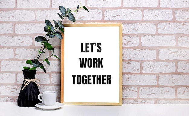 On a white table against a light brick wall a branch of eucalyptus in a dark vase a white cup and a light wooden frame with the text LET IS WORK TOGETHER Home office interior