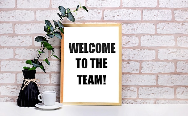 On a white table against a light brick wall a branch of eucalyptus in a dark vase a white cup and a light wooden frame with the text WELCOME TO THE TEAM Home office interior