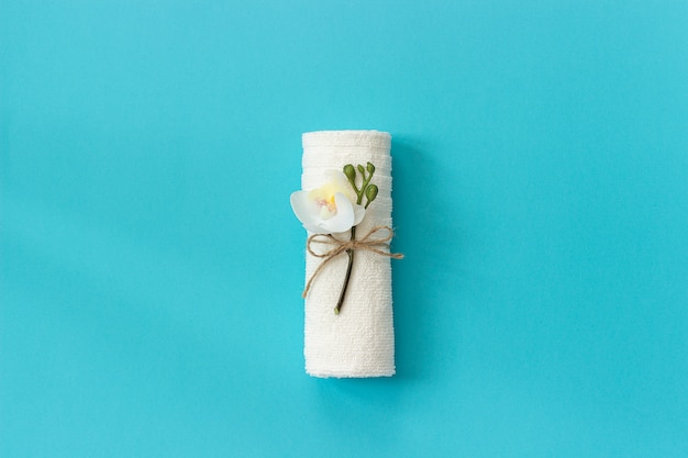 White towel roll tied with rope with sprig of orchid flower on blue paper background.