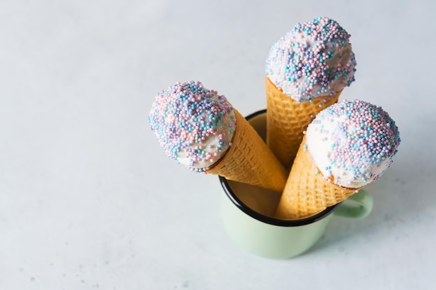 White vanilla ice cream scoop in waffle cone with sprinkles in cup on light grey background