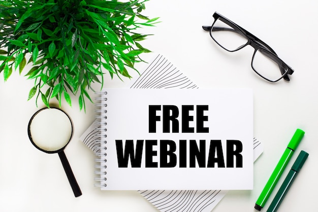 On a white wall lies a notebook with the word FREE WEBINAR, glasses, a magnifying glass, green markers and a green plant