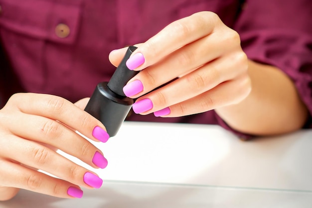 White woman holding nail polish black bottle with painted pink nails close up.