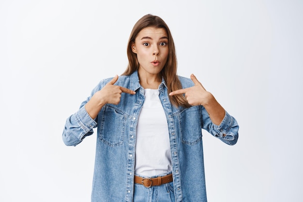 Who me. Surprised blond girl pointing at herself with disbelief, staring startled, being chosen or named, standing on white