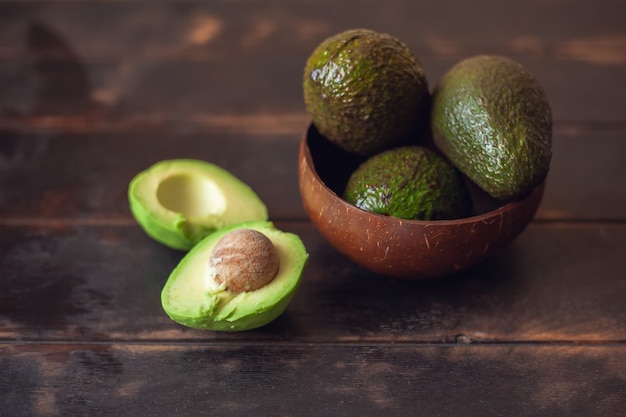 Whole avocado fruits in a bowl made from coconut shells and cut in half lie next to it on a dark wooden background.