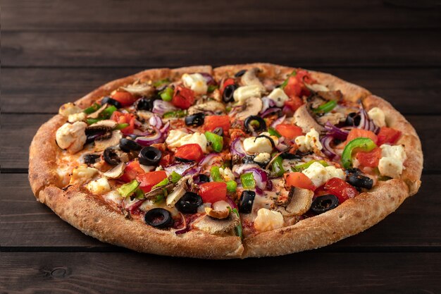 Whole fresh round pizza with chicken meat, vegetables, mushrooms and cheese close-up on a wooden brown table. 