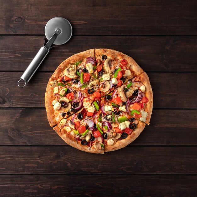 Whole fresh round pizza with chicken meat, vegetables, mushrooms, cheese and knife top view on a wooden brown table. 