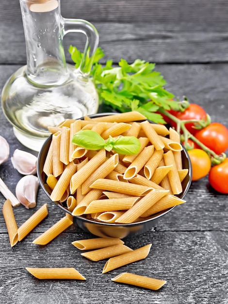 Whole grain flour penne pasta in a bowl tomatoes garlic vegetable oil in a glass decanter and parsley on dark wooden board background