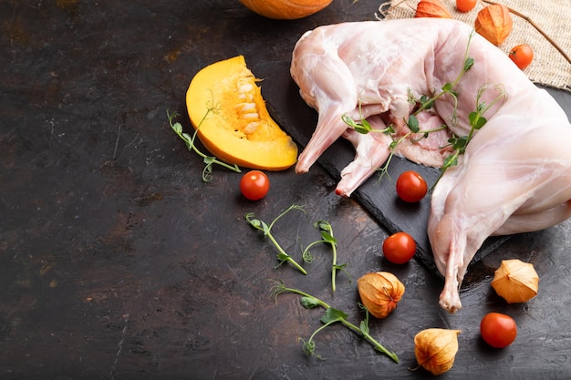 Whole raw rabbit with pumpkin, tomatoes, pea sprouts on a black concrete background and linen textile. Side view, copy space.