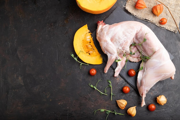 Whole raw rabbit with pumpkin, tomatoes, pea sprouts on a black concrete background and linen textile. Top view, flat lay, copy space.