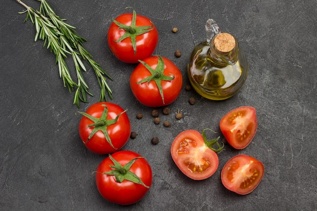 Whole tomato and chopped tomato wedges, allspice on table.  Bottle of olive oil and sprig of rosemary.  Black background. Top view.