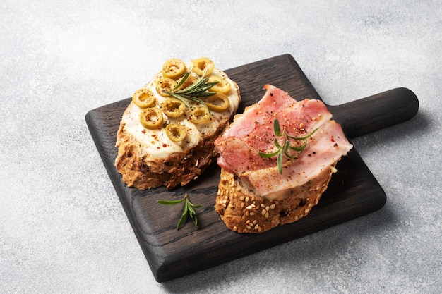 Wholegrain bread sandwiches with cream cheese, bacon and olives on a wooden cutting Board. A glass of white wine.