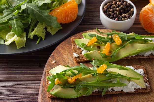 Wholegrain crisp bread with cheese, avocado, fresh leaves and tangerine on wooden backdrop