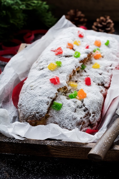 Wholegrain Stollen with raisins and powdered sugar on a linen napkin with a sieve, red ribbon over the old dark concrete