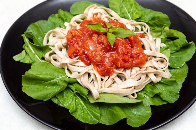 Wholemeal pasta pasta with green leaves of arugula, red sauce and balsamic vinegar on black plate and white marble background