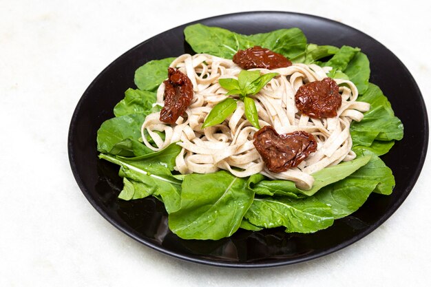 Wholemeal pasta with green leaves of arugula and dried tomato on black plate and white marble