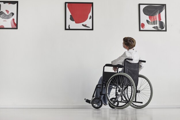 Photo wide angle portrait of young woman using wheelchair looking at paintings in modern art gallery, 