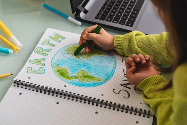 Wide angle view of child girl draws planet earth with wax colors on school notebook for Earth day Little activist girl writes the message Save the Planet Protection of environment global warming