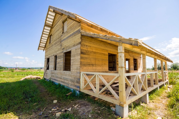 Wide angle view of unfinished wooden ecological traditional cottage of natural lumber materials with steep roof and porch under construction in green neighborhood on blue sky copy space background.