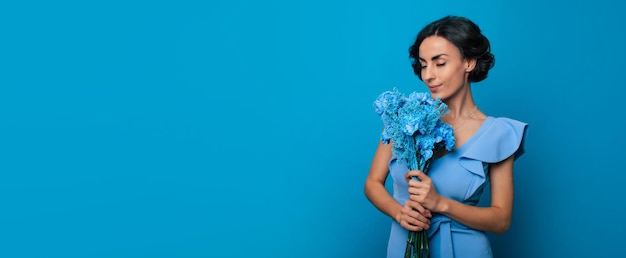 Wide banner photo of young gorgeous woman in a bright blue dress is looking in the camera with a big smile, holding a bunch of blue flowers in her hands