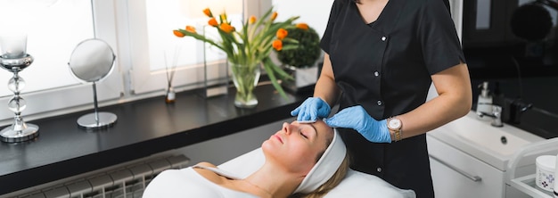 Photo wide indoor shot of unrecognizable female cosmetician in black uniform and blue protective gloves using cosmetic swaps on the face of her caucasian female client spa massage