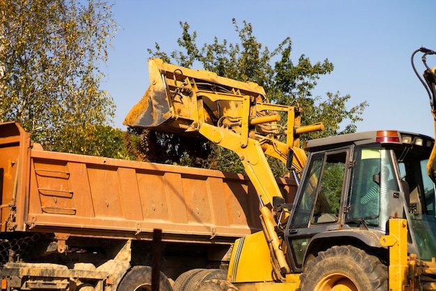 A wide metal excavator bucket digs sand or clay and loads it into a dump truck at a construction site.