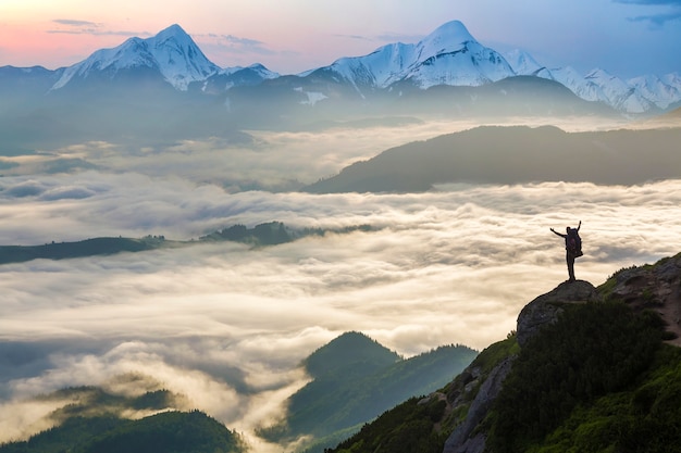 Wide mountain panorama. Small silhouette of tourist with backpack on rocky mountain slope with raised hands over valley covered with white puffy clouds. Beauty of nature, tourism and traveling concept