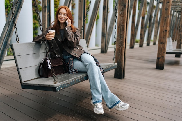 Photo wide shot of pretty young woman with long red hair talking on smartphone swinging on city swings