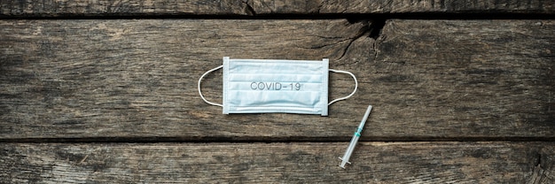 Wide view image of a medical protective mask with Covid 19 sign and  an injection placed over rustic wooden background.