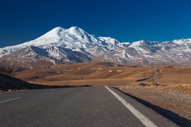 Winding road leads through hills to snowcovered rocky mountains against clear sky Caucasus Russia