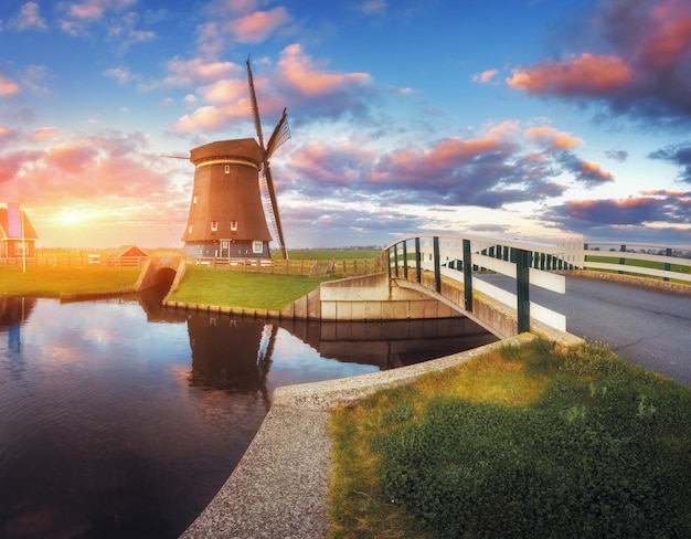 Windmill and bridge near the water canal at sunrise in Netherlands. Traditional dutch windmill against colorful sky in dusk. Spring landscape in Holland. Sunny morning. Cloudy sky reflected in water