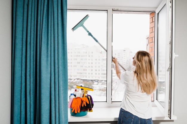 Windows cleaning service Housewife is doing chores and washing windows indoor in apartment Woman cleaner with mop is cleaning window at home