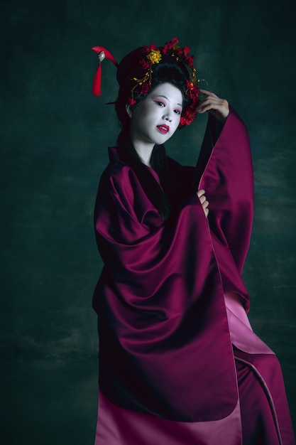 Wine color. Young japanese woman as geisha isolated on dark green background. Retro style, comparison of eras concept. Beautiful female model like bright historical character, old-fashioned.