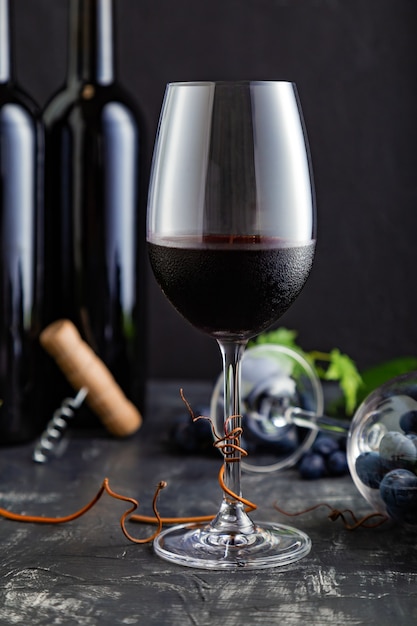 Wine glass with red wine. Red Wine bottles, grape bunches with leaves and vines on dark rustic concrete background. Wine composition on black stone table.