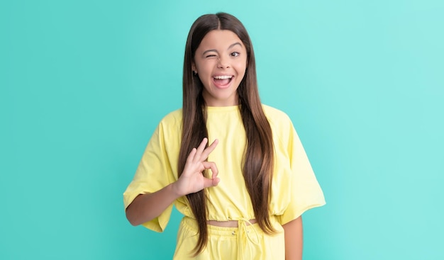 Winking kid with long hair show ok gesture on blue background with copy space teen