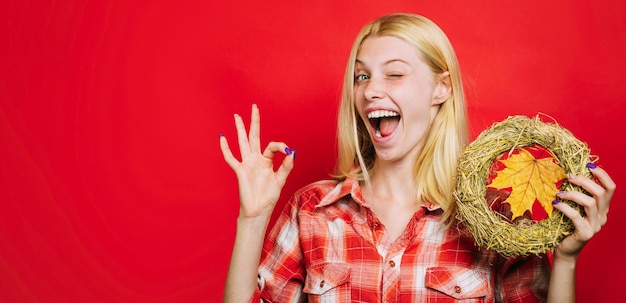 Winking woman in plaid shirt with decorative autumn wreath and maple leaf showing ok sign