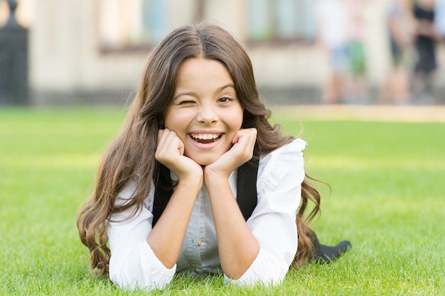 Winking to you. School break for rest. Adorable pupil. Girl kid laying lawn. Girl school uniform enjoy relax. Importance of relaxation. Little schoolgirl. Relax at school yard. Kid relaxing outdoors.