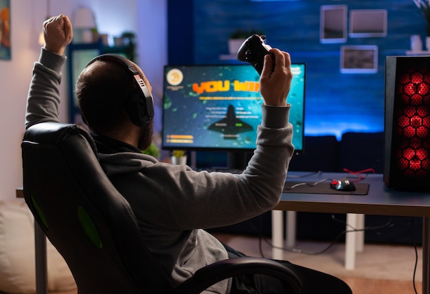 Winner gamer sitting on gaming chair at desk and playing space shooter video games with controller. Man streaming online videogames for esport tournament in room with neon lights