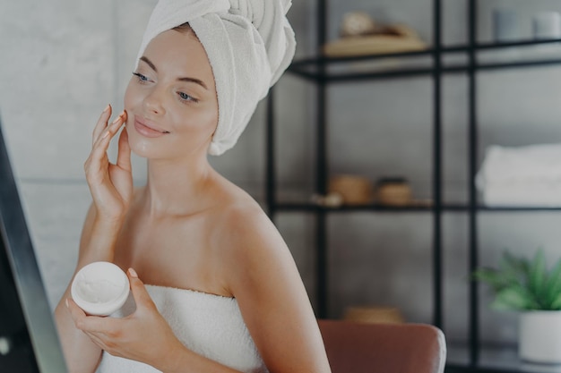 Winsome pleased young European woman puts cream on face looks at her reflection in mirror wrapped in bath towel has anti aging procedure after showering for healthy skin Skin care concept