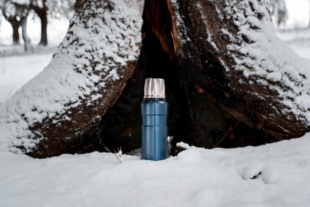 Winter camping in the forest concept. Thermos standing on the background of an old tree. Travel Vacuum Flask