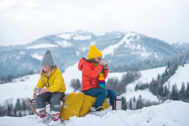 Winter children travellers enjoy coffee time with scenery view of the snowy mountain background. Kids in snow drink tea.