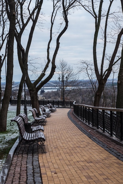 Winter cloudy day in the city park. View on the river, benches and light snow. City landscape view.