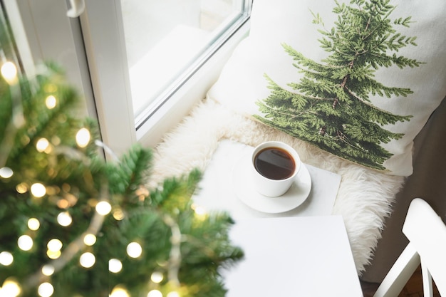 Winter coffee on window sill with Christmas tree and pillow for cozy reading