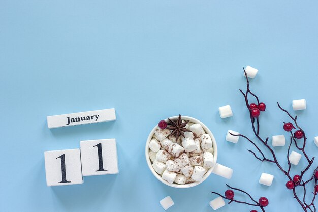 Winter composition. White wooden calendar cubes. Data January 11. Cup of cocoa, marshmallows and decorative branch with red berries on blue background Top view Flat lay Copy space