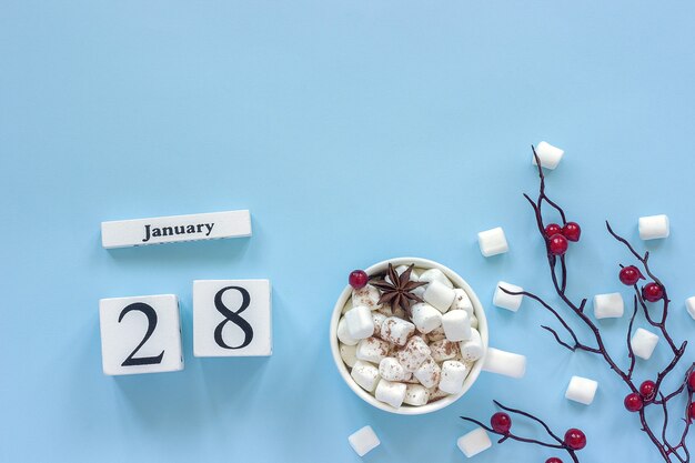 Winter composition. White wooden calendar cubes. Data January 28. Cup of cocoa, marshmallows and decorative branch with red berries on blue background Top view Flat lay Copy space